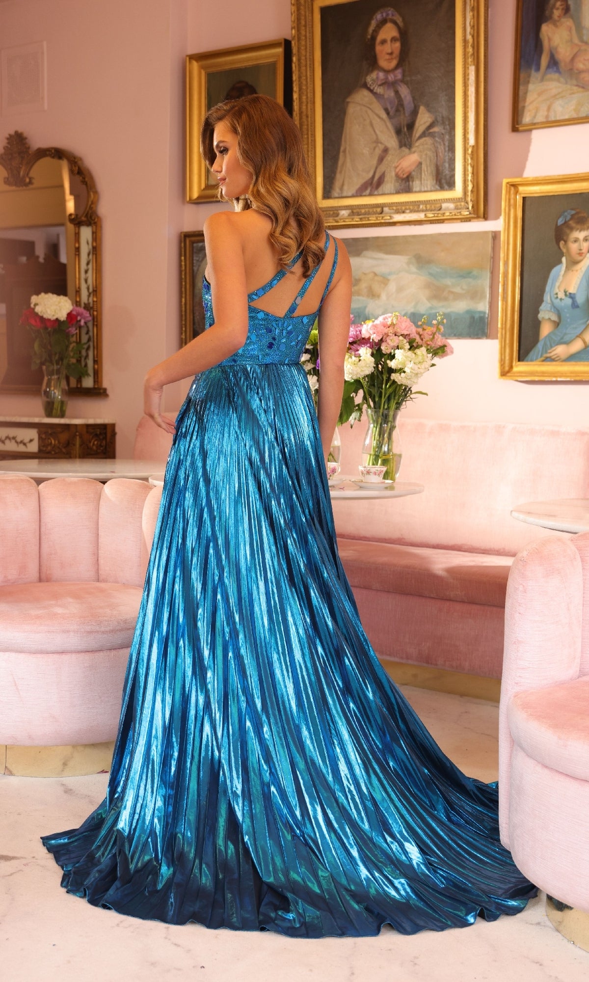A woman wearing the Ava Presley 40105 Long Metallic Pleated Prom Gown stands in an elegant room with pink sofas, flowers, and framed portraits on the wall.